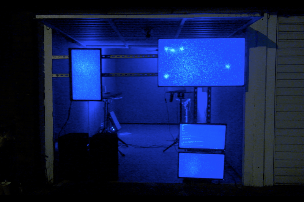 A garage is filled with blue light. Screens suspended on a metal track are hung in the open doorway to the garage. Each of the four screens are blue.