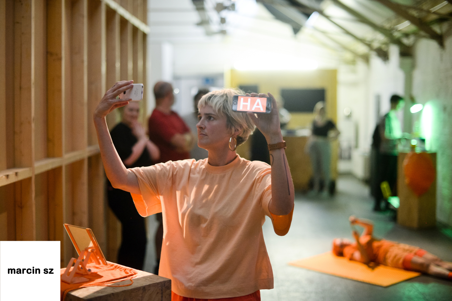 Emily W, dressed in full orange stands with arms raised holding two mobile phones up. They look at one screen whilst the other faces the camera. On it has big white letters reading 'HA' on an orange background.