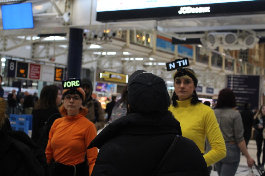 Two performers are wearing phones attached to their head. They are both looking into the camera wearing yellow and orange.