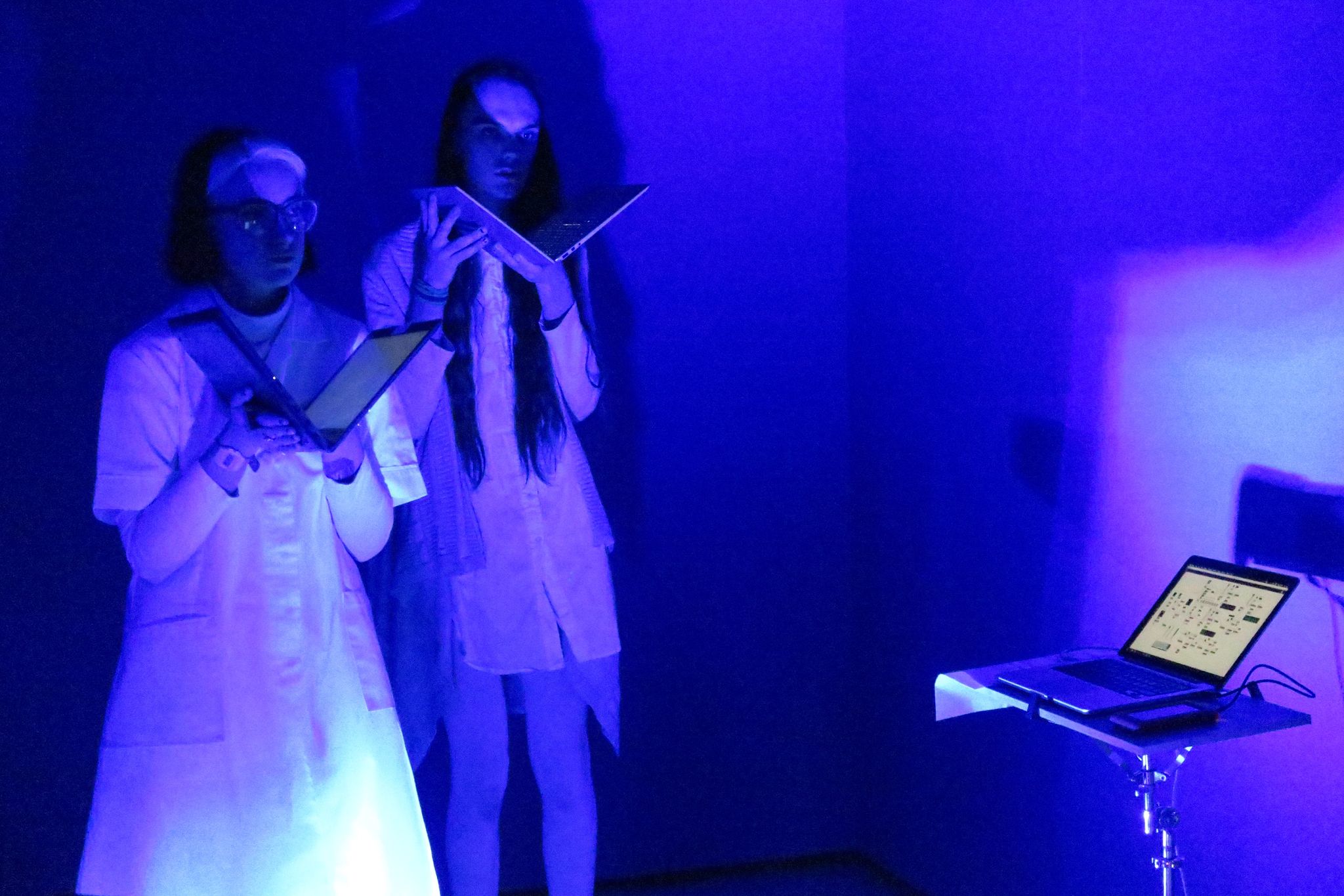 Georgina and Rubie stand dressed in white in a blue room. They each hold a laptop open like a book below their faces.