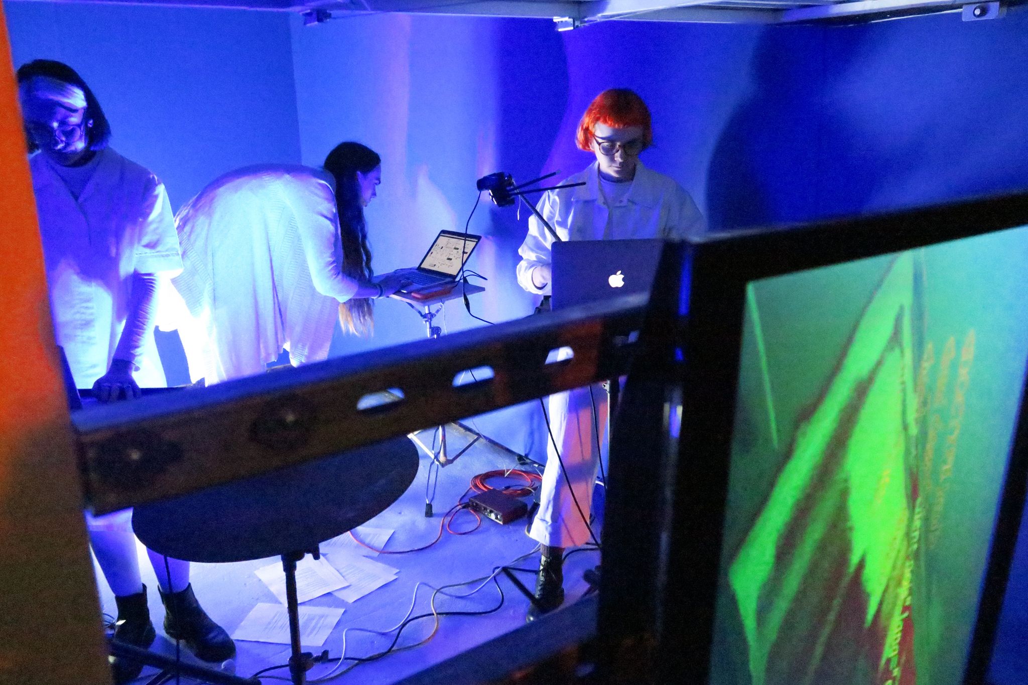 Computer screens and tvs are hung on a metal railing. Behind are the three performers stood dressed in white looking at laptops in a blue painted garage. Cables and papers cover the floor.