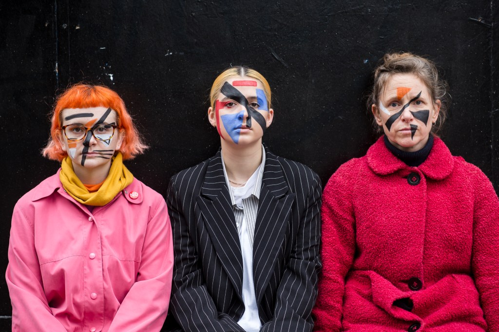 Emily, Evie and Anna sit in front of a black background facing the camera with straight faces. Each of them have bright blocky shapes painted on their faces in black, white, blue and red.