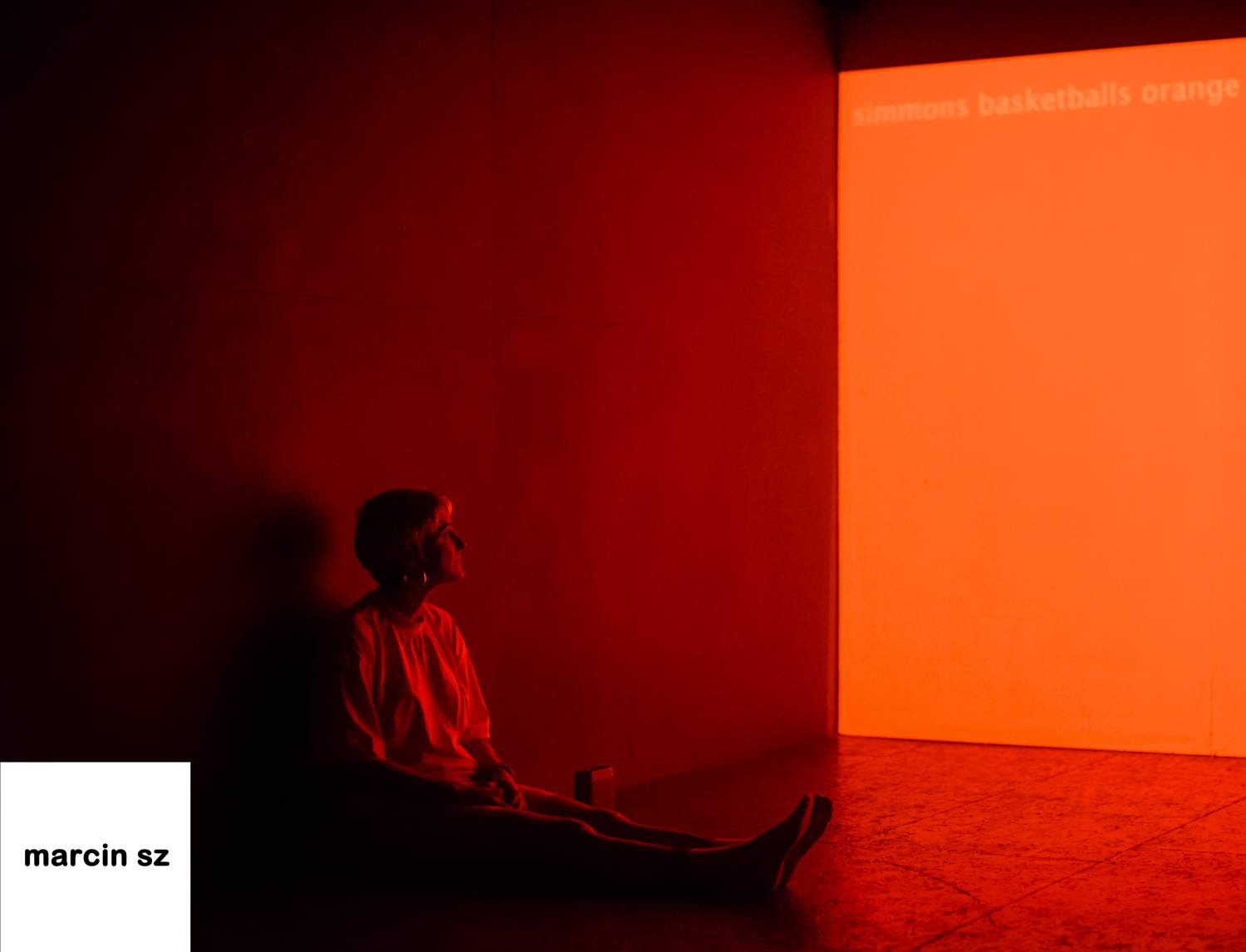 Emily W, dressed in orange sits up against a wall in a gallery watching a full orange projection. It glows into the space.
