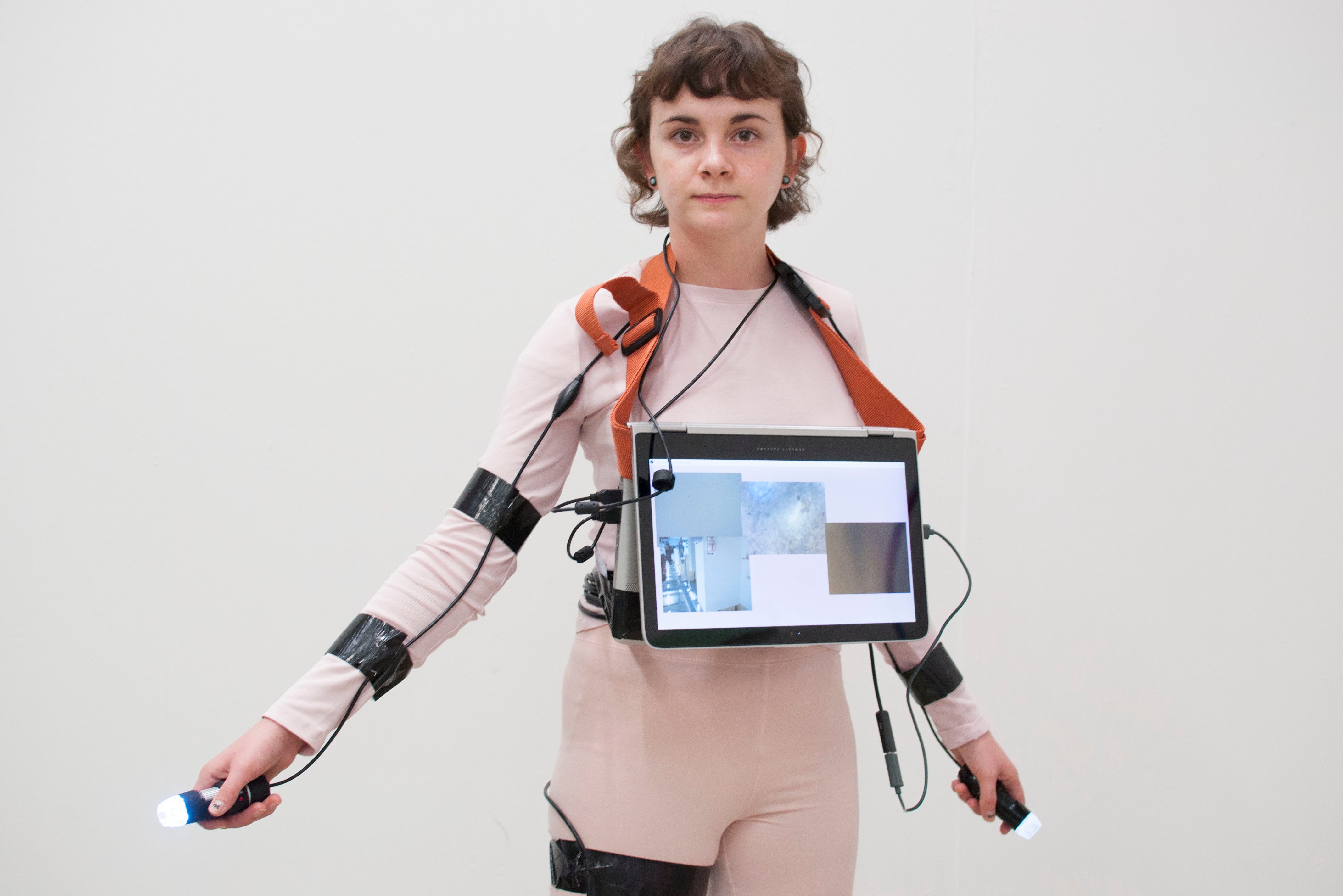 Emily stands infront of a white wall with her arms lowered but out to the side slightly. She wears a screen hung from her neck that covers her chest. She wears pink clothes with black tape and cables that cover her linbs. In both hands are small camera devices.