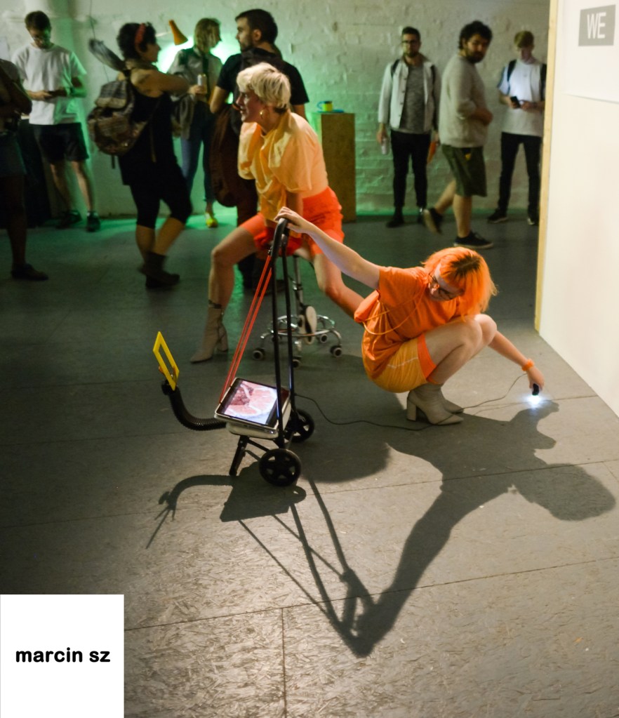 Emily and Emily, dressed in orange are low to the ground, one on a stool and the other crouched down. A laptop folded on a wheelie stand is central to the image.