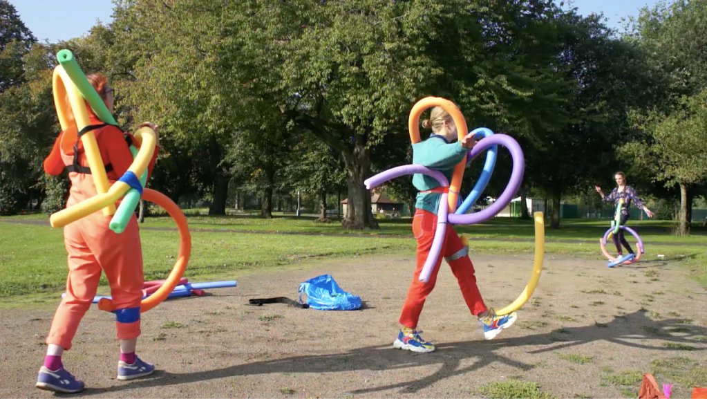 In a grassy park area surrounded by thick green trees, three people stand wearing colourful clothing and have multiple swimming noodles in various colours attached to their bodies. They curve out away from the bodies, creating limb-like extensions.