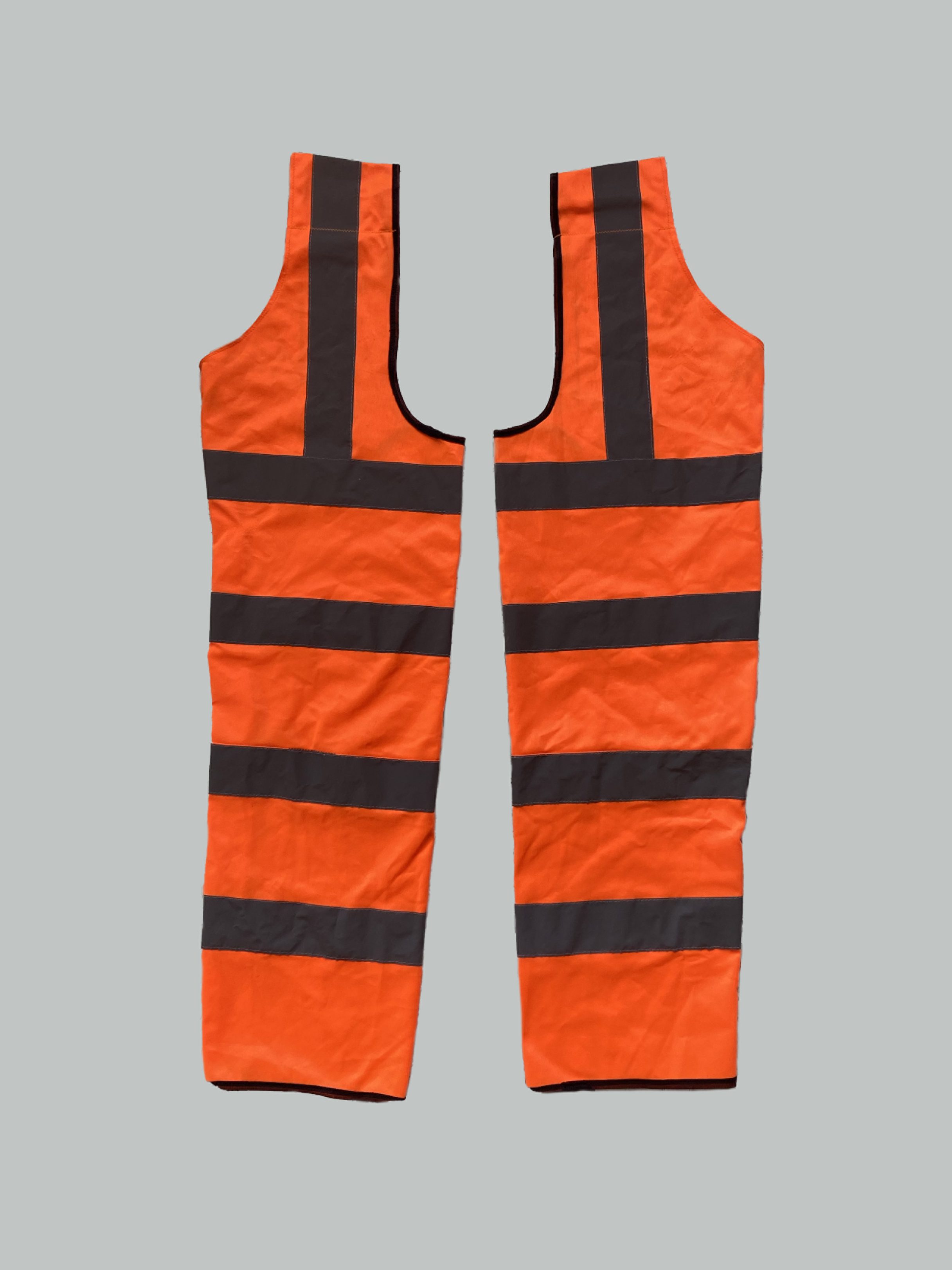 On a soft grey background in portrait format are a pair of high-vis full chaps (also recognised as trousers with the bottom and crotch exposed). They are orange striped high-vis fabric with reflective horizontal strips.