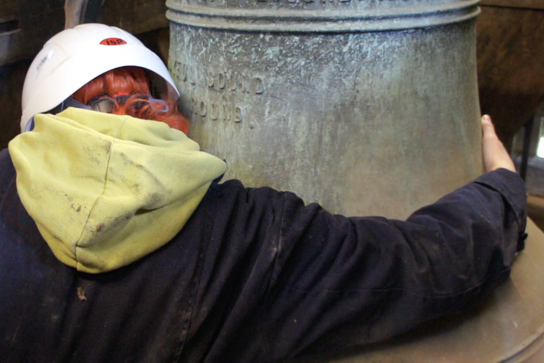 Emily is crouched facing away from the camera, hugging a large church bell. Her arms don't reach all the way around and her orange hair pokes out the bottom of her white hard hat.