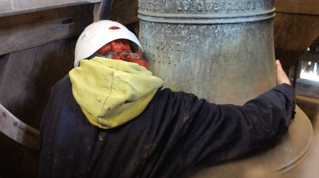 Emily is crouched facing away from the camera, hugging a large church bell. Her arms don't reach all the way around and her orange hair pokes out the bottom of her white hard hat.