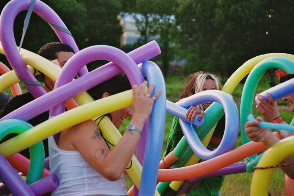 Photograph close up of a group of people covered in various swimming noodles in a grassy park area.