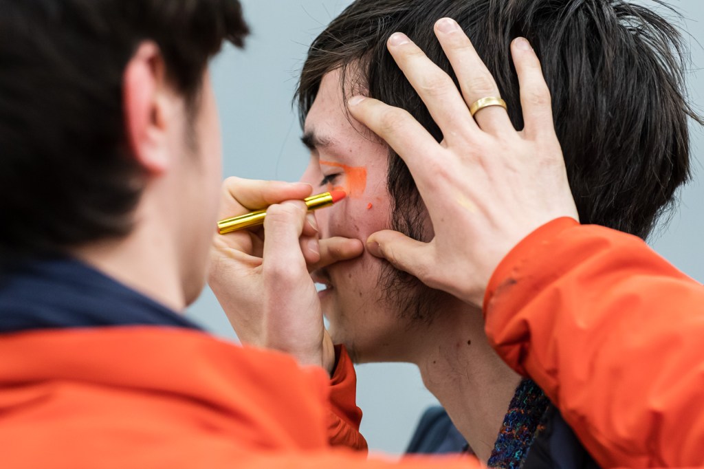 Two white men are close to the camera. One draws on the other's face in orange face crayon.