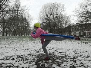 A GIF of Emily moving a blue swimming noodle that is attached to one of her feet. She stands on one leg and moves the noodle low around her feet, she then extends and lifts her leg before pointing her toe into the mud and steching backwards with the noodle. She is in a muddy and snowy park area.