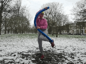 A GIF of Emily moving a blue swimming noodle that is attached to one of her feet. She stands on one leg and moves the noodle low around her feet, she then extends and lifts her leg before pointing her toe into the mud and steching backwards with the noodle. She is in a muddy and snowy park area.