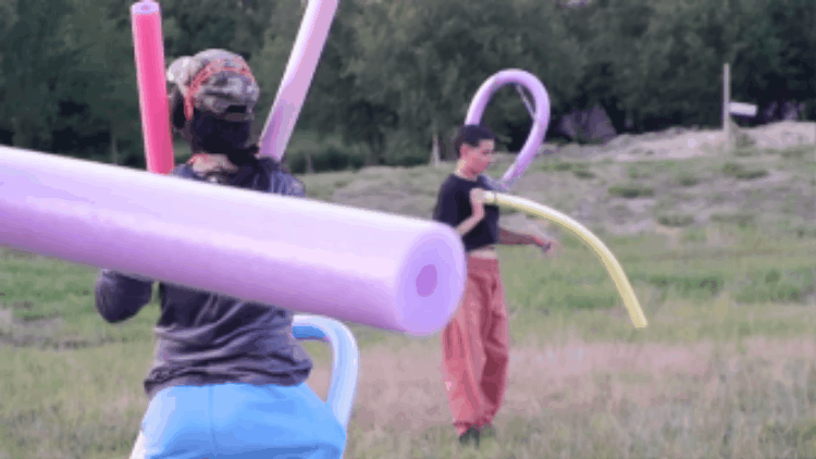 Short animated GIF. Two people in a field are wearing multiple noodles curled and bent in different ways. They move slightly and bend one of the noodles. A purple noodle enters the left of the frame blocking some of the view.