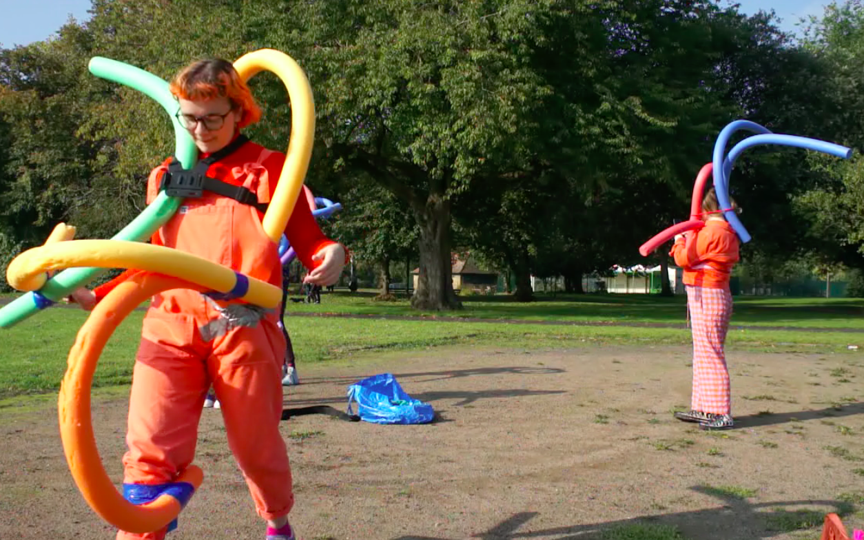 In a bright sunny park, two people in bright clothes are wearing multiple swimming noodles strapped to them in different ways. They curl up and away from the body.