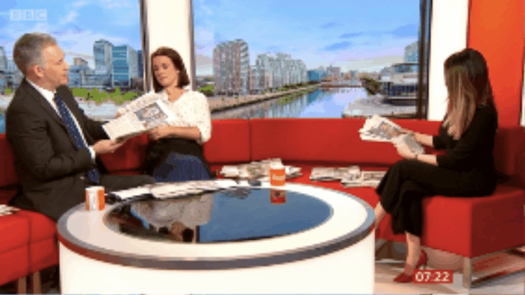 GIF image plays of three TV presenters on a sofa holding up newspapers to camera, the newspaper spread has a mosaic of headshots of the Dazzle Club members with paint CV Dazzle faces.